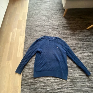 Marinblå kabelstickad tröja Gant - Kabelstickad marinblå tröja från Gant med rund hals och diskret broderad logga på bröstet. Tröjan har långa ärmar och ribbade muddar vid ärmslut och nederkant. Perfekt för dig som gillar klassisk och stilren look. 