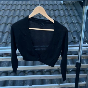 Svart croppad kofta från H&M - Svart, tunn och croppad kofta från H&M med lång ärm och djup v-ringning. Koftan har en enkel knapp framtill och är gjord i ett lätt, stickat material som känns luftigt. Perfekt att slänga över ett linne eller klänning för en chill look.