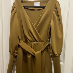 Olivgrön omlottklänning från ASOS - Snygg olivgrön omlottklänning från ASOS DESIGN med långa puffärmar och markerad midja med bälte. Klänningen har v-ringning och stilren design som ger en cool och trendig look.