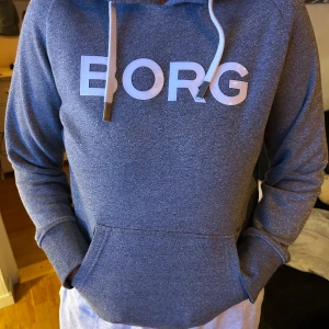 Grå hoodie från BORG med tryck - Säljer en snygg grå hoodie från BORG med stor vit logga framtill. Tröjan har huva med vita snören och en klassisk känguruficka. Perfekt för chill dagar eller när du vill ha en sportig look.