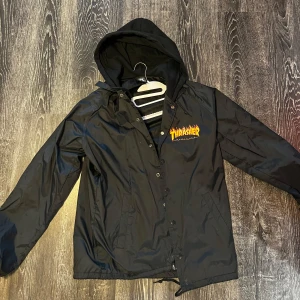 Svart Thrasher vindjacka med huva - Svart vindjacka från Thrasher med klassisk logga i gult och rött på bröstet. Jackan har huva, knappar framtill och dragsko nedtill. Perfekt för dig som gillar streetwear och vill ha en stilren jacka med attityd.