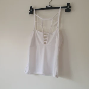 Vit linnetopp med coola banddetaljer strl M - Säljer en vit linnetopp med smala axelband och unika banddetaljer både fram och bak. Toppen har en djup v-ringning med horisontella band och är tillverkad i ett stretchigt, mjukt syntetmaterial. Perfekt för dig som gillar en stilren men edgy look.  Helt skick dock små obehandlade fläckar vid halsen
