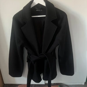Svart kappa från Gina Tricot med bälte - Stilren svart kappa från Gina Tricot med klassisk krage och knytbälte i midjan. Jackan har lång ärm och en rak, något loose passform som ger en snygg siluett. Perfekt för dig som gillar minimalistisk och tidlös stil.