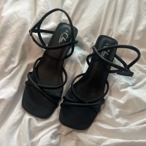 Svarta sandaler med klack och remmar - Stilrena svarta klackar med fyrkantig tå, tunna remmar och klack. Snygg minimalistisk design med öppen tå och justerbar ankelrem. Perfekta för dig som gillar en clean look med lite edge.