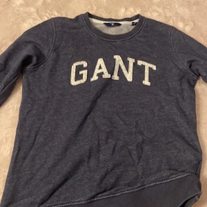 Mörkblå sweatshirt från GANT - Mörkblå sweatshirt från GANT med klassisk rund hals och stora vita bokstäver framtill. Tröjan har långa ärmar och ribbade muddar vid ärmslut och nederkant. Perfekt för en chill och stilren look.