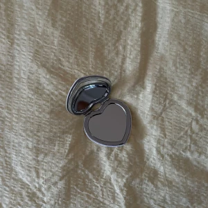 Hjärformad sminkspegel med kaninmotiv - Supersöt hjärtformad sminkspegel i silverfärgad metall med vit baksida. Ena sidan har ett gulligt mönster med kaniner i tekoppar och rosa rosetter. Perfekt storlek för väskan och smidigt att ta med sig överallt.