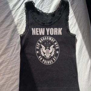 Svart linne med spets och New York-tryck - Svart ribbad linnetopp från Brandy Melville med vit text 'NEW YORK' och emblemtryck framtill. Spetsdetaljer längs halsringning