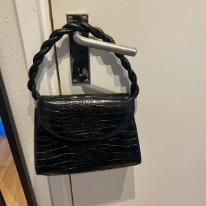 Svart handväska med kedja - Snygg svart handväska i croco-imitation med flätat handtag och guldfärgad kedja som axelrem. Väskan har ett stilrent lock och detaljer i guld. Den är lite sliten men det går enkelt att fixa, jag har bara inte haft tid eller ork till det, därför säljer jag den så billigt. 
