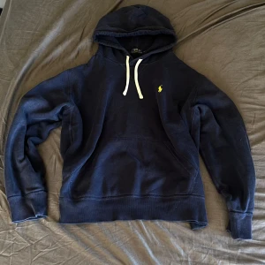 Marinblå hoodie från Polo Ralph Lauren - Snygg marinblå hoodie från Polo Ralph Lauren med klassisk gul logga på bröstet. Tröjan har huva med vita snören och en stor magficka. Närmare small än medium, passar nån som är runt 175 kanske