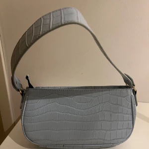 Ljusblå axelväska i crocopräglat skinn - Säljer en ljusblå axelväska med crocopräglat mönster och bred axelrem. Väskan har guldfärgade detaljer och stängs med dragkedja. Perfekt storlek för det viktigaste och ger en trendig touch till din outfit.