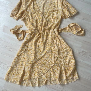 Gul blommig omlottklänning med volang - Superfin gul kortklänning med blommigt mönster, omlottmodell och knytband i midjan. Klänningen har korta ärmar och volangkant nertill, perfekt för sommarens varma dagar. Lätt och luftig känsla, passar dig som gillar en söt och avslappnad stil.