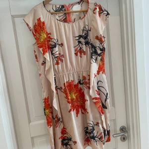Blommig kortklänning från InWear - Superfin kortklänning från InWear i ljusbeige med stora blommiga mönster i orange nyanser, lite gult, lite grått och svart. Klänningen har volangdetaljer längs sidorna, rund halsringning och dragkedja bak. Perfekt för dig som gillar färg och vill sticka ut lite extra, men som ändå vill ha en neutral honungsbeige bas. 