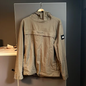 Vindjacka från Weekend Offender - Stilren beige vindjacka från Weekend Offender med huva och logotyp på ärmen. Jackan har dragsko nertill och diskreta fickor. Perfekt för dig som vill ha en clean look med streetkänsla. Materialet är lätt och passar bra till vår och höst.