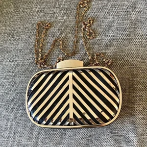 Svart och guld randig kuvertväska - Snygg kuvertväska i svart skinn med guldiga metallränder i ett coolt mönster. Väskan har en hård, oval form och en kedja i guld som axelrem. 