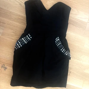 Svart kort klänning med detaljer - Snygg svart mini klänning med hjärtformad urringning och axelbandslös design. Klänningen har glansiga silver stenar vid fickorna och en dragkedja med hjärtformad dragkedja på ryggen