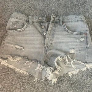 Grå slitna jeansshorts - Snygga grå jeansshorts med hög midja och slitna detaljer. Shortsen har fransig kant nertill, klassiska fem fickor och knappar framtill. Perfekta för en avslappnad och trendig look under sommaren.