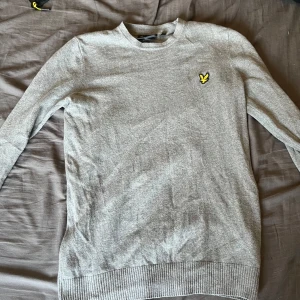 Grå stickad tröja från Lyle & Scott - Snygg grå stickad tröja från Lyle & Scott med klassisk rund hals och deras ikoniska gula fågel-logga broderad på bröstet. Tröjan har långa ärmar och ribbade muddar för en clean look. Perfekt för lager-på-lager under hösten och vintern. Pris ej hugget i sten vid snabb affär. hör av dig vid minsta fundering!