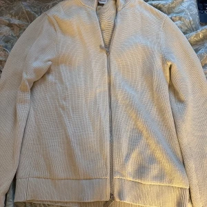 Beige kofta från Selected Homme - Snygg beige kofta från Selected Homme i stickad bomull. Modellen har hel dragkedja framtill, ribbade muddar och krage. Perfekt för lager-på-lager och passar till många olika stilar. Den är använd cirka 2 gånger och är i nyskick, nypris är 800 kr