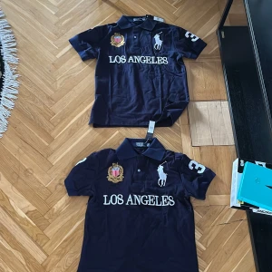 Mörkblå pikétröja Polo Ralph Lauren - Snygg mörkblå pikétröja från Polo Ralph Lauren med broderad logga, stort tryck 'LOS ANGELES' och siffra 3 på ärm och rygg. Klassisk krage, knappar framtill och detaljer i vitt och guld. Perfekt för en sportig och stilren look.