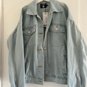Ljusblå jeansjacka från Boohoo - Säljer en snygg ljusblå jeansjacka från Boohoo med klassisk krage, bröstfickor med knappar och raka sömmar. Jackan har en avslappnad passform och är perfekt att slänga över en hoodie eller t-shirt. Stängs med silvriga knappar framtill.