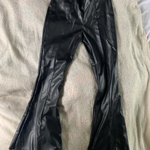 Svarta bootcut byxor i skinnimitation - Säljer ett par svarta bootcut byxor från Divided i skinnimitation. Byxorna har medelhög midja och en snygg glansig finish som ger en edgy vibe. Perfekta för dig som vill sticka ut med en trendig och cool look. Endast använd en gång