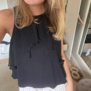 Svart ärmlös blus med volang - Säljer en svart ärmlös blus med snygga volangdetaljer framtill och på sidorna. Blusen har rund halsringning och ett lätt, flowy tyg som ger en luftig känsla. Perfekt att styla med jeans eller kjol för en trendig look.