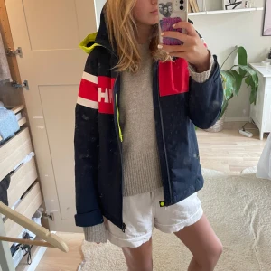 Helly Hansen vindjacka marinblå/röd - Snygg vindjacka från Helly Hansen i marinblått med röda och vita detaljer. Jackan har dragkedja framtill, huva och två fickor med neonfärgade dragkedjor. Använd en säsong så bra skick! Skicka privat för fler bilder/ frågor 