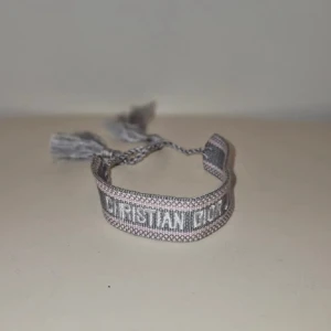 Grått armband från Christian Dior - Snyggt vävt armband från Christian Dior i grå och silverton med tydlig logga broderad framtill. Armbandet har justerbar knytning med tofsar i ändarna för en cool och avslappnad look. Perfekt accessoar för att lyfta din outfit.
