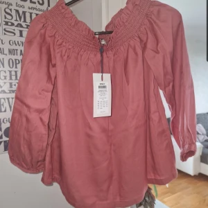 Rosa offshoulder blus från ONLY - Supersöt rosa offshoulder blus från ONLY med smockad resår upptill och 3/4-ärmar. Blusen har en luftig passform och är gjord i mjuk bomull, perfekt för vår och sommar. Enkel att styla med både jeans och kjol.