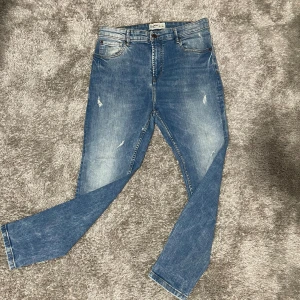 Blå jeans från Pull&Bear - Snygga blå jeans från Pull&Bear i storlek 44. Modellen har klassisk femficksdesign, ljus tvätt med slitningar och smal passform. Jeansen är tillverkade i bomull och har en modern look med lätt slitna detaljer framtill.