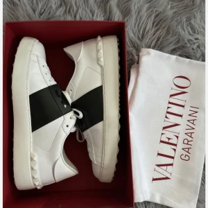 Valentino Garavani vita sneakers - Snygga vita sneakers från Valentino Garavani med bred svart rem över mitten och ikoniska nitar på hälen. Skorna har rund tå, platt sula och är tillverkade i skinn. Perfekta för dig som gillar exklusiv streetstyle.