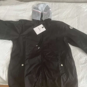 Svart vindjacka från Moncler med huva - Svart vindjacka från Moncler med klassisk logga på ärmen och dragkedja framtill. Jackan har en tunn, genomskinlig huva med röd och vit rand, samt praktiska fickor med tryckknappar. Perfekt för dig som vill ha en stilren och sportig look.
