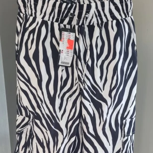Zebramönstrade cargopants med snörning - Säljer ett par svarta och vita cargopants med zebramönster. Byxorna har elastisk midja med snörning och stora fickor på sidorna. Materialet är lätt och känns skönt mot huden, perfekt för dig som vill sticka ut med en cool look.