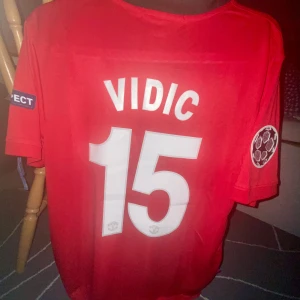 Manchester United Vidic 15 Nike tröja - Manchester Uniteds officiella matchtröja med Vidic 15 på ryggen. Röd med svart V-detalj över bröstet, klubbmärke, Nike-logga och AIG sponsortryck. Champions League och UEFA Respect-patchar på ärmarna. Tillverkad i lätt och ventilerande polyester. Det är inte slutpriset på tröjan så om du är intresserad skicka ett bud och vi kan diskutera den