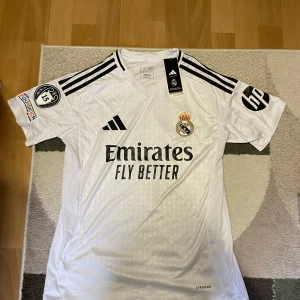 Real Madrid Vini Jr. 7 Adidas tröja M - Real Madrid matchtröja i vitt med svarta detaljer, Adidas-märke och klubbmärke på bröstet. VINI JR. och nummer 7 på ryggen. Kortärmad modell i lätt funktionsmaterial, slim fit och klassiska svarta ränder på axlarna. Snygg V-ringning och officiella sponsortryck.