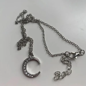 Halsband - Snyggt halsband i silverfärg med en glittrig måneberlock. Kedjan är tunn och elegant, perfekt för att lägga till en touch av stjärnglans till din outfit. Månen är dekorerad med små gnistrande stenar som fångar ljuset vackert. 