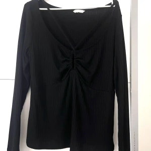 Svart ribbad långärmad topp  - Svart långärmad topp med ribbad struktur och snygg snörning framtill vid bysten. Djup v-ringning och smala band som korsas över bröstet.