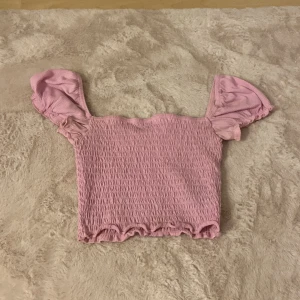 Rosa smockad topp från Divided XS - Supersöt rosa smockad topp från Divided med puffiga korta ärmar och volangkant nertill. Toppen har en croppad passform och är perfekt för dig som gillar en feminin och trendig stil. Mjuk och stretchig i materialet för extra komfort.