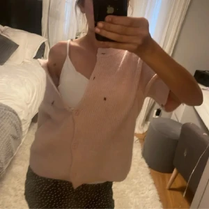 Ljusrosa stickad kofta med knappar - Söt ljusrosa stickad kofta med korta ärmar och knappar framtill. Koftan har en avslappnad passform och är perfekt att slänga över ett linne. Enkel och stilren design som passar till många outfits.