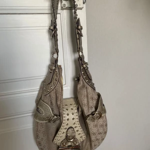 Beige axelväska från Guess med nitar - Säljer en beige axelväska från Guess med logomönster, detaljer i ljus skinnimitation med krokoprägling och silverfärgade nitar. Den är i jättefint skick, men har tappat några stenar som man ser på guess loggan. Nypris ligger runt 2000kr, så säljes för 900!