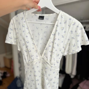 Vit blommig topp Gina Tricot - Supersöt vit kortklänning från Gina Tricot med ljuslila blommönster. Klänningen har v-ringning, korta ärmar och knyt i midjan. Perfekt för sommarens alla tillfällen och ger en fräsch och romantisk vibe.