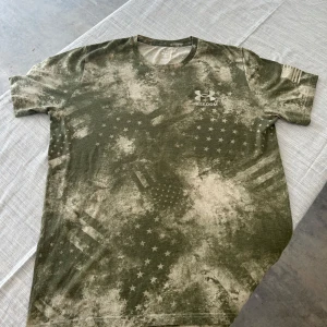 Grön camo t-shirt från Under Armour - Snygg grön t-shirt från Under Armour Freedom-kollektionen med camouflagemönster och stjärnor. Tryckt amerikansk flagga på ärmen och stort Under Armour-logo på bröstet. Modellen är loose fit och har korta ärmar. Perfekt för dig som gillar streetwear och sportig stil.