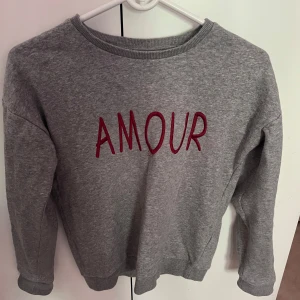 Grå sweatshirt med AMOUR text från Name It - Grå sweatshirt från Name It med röd broderad text 'AMOUR' på bröstet och liten svart text 'Fait avec amour' i nacken. Mjuk bomullskänsla, rund hals och långa ärmar. Perfekt för dig som gillar enkel men snygg och lite semester stil. Knappt använt 
