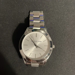 Silverfärgad klocka från Michael Kors - Snygg och stilren klocka från Michael Kors i silverfärg. Den har en rund urtavla med minimalistiska markeringar och ett armband i rostfritt stål. Perfekt för en elegant look. OBS! saknar sprint som på sista bilden. En liten sprint som ska sättas fast i det lilla svarta hålet. Går att köpa billigt. Klockan har en enstaka repa som knappast syns vid vanlig användning. Går att minska i storleken med lite erfarenhet. Pris kan diskuteras 