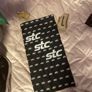 Snygg STC sport halsduk - Snygg svart halsduk med vit stc-logga över hela tyget. Perfekt för att ge en sportig touch till din outfit. Halsduken är lätt och smidig, vilket gör den enkel att bära med sig