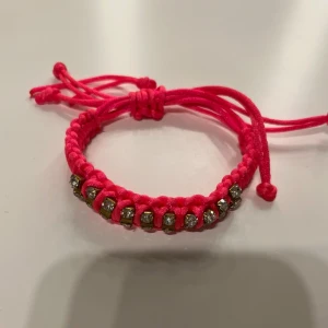 Rosa armband med glittriga detaljer - Snyggt rosa armband med justerbar knytning och glittriga pärlor. Perfekt för att ge en färgklick till din outfit. Armbandet har en flätad design och är lätt att anpassa i storlek.