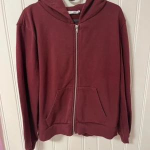 Vinröd hoodie med dragkedja - Säljer en vinröd hoodie med dragkedja framtill. Den har långa ärmar och en praktisk huva. Perfekt för kyligare dagar och avslappnade stunder. Hör av dig vid intresse!
