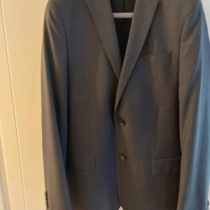 Grå kavaj - Elegant grå kavaj med klassisk design och två knappar framtill. Perfekt för formella tillfällen eller en stilren vardagslook. Kavajen har en bröstficka och två fickor nedtill.
