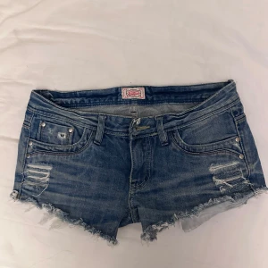 Blå jeansshorts med fransar - Snygga blå jeansshorts från SoCal med slitna detaljer och fransar vid benen. Shortsen har en låg midja och klassisk femficksdesign med nitar på fickorna. Perfekta för en avslappnad stil.