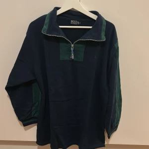 Mörkblå tröja med dragkedja från Polo - Säljer en mörkblå tröja från Polo med gröna detaljer och dragkedja framtill. Tröjan har långa ärmar och en avslappnad passform, perfekt för kyligare dagar. Den klassiska designen gör den lätt att matcha med olika stilar.